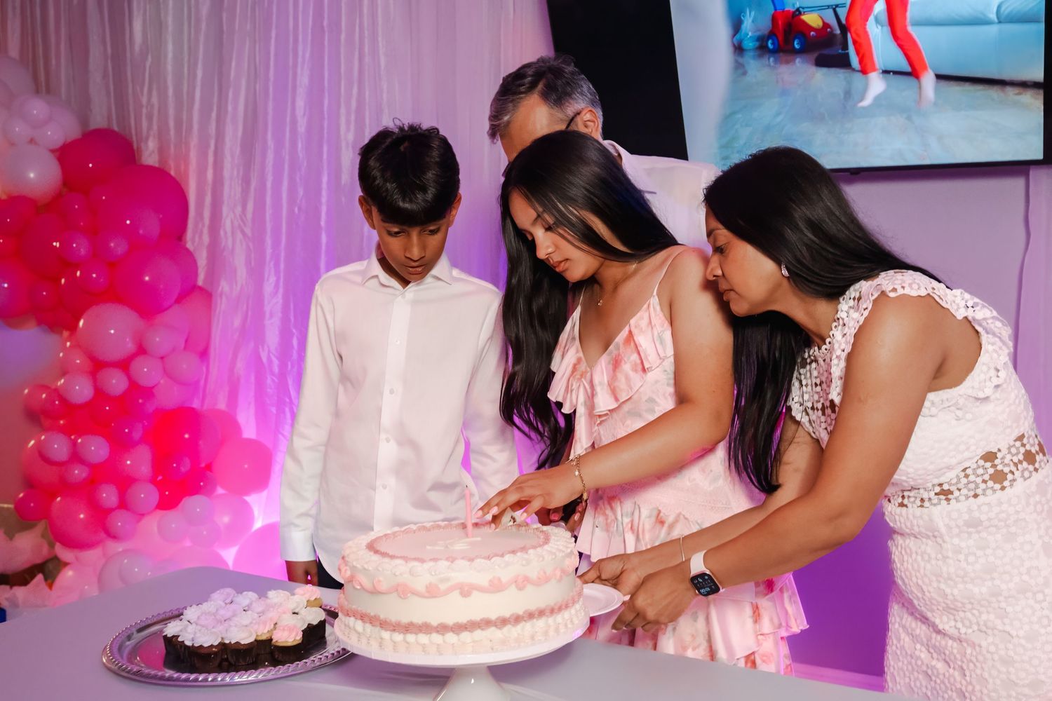 Teen girl cutting her Sweet 16 cake with help from family at a pink balloon-themed party.