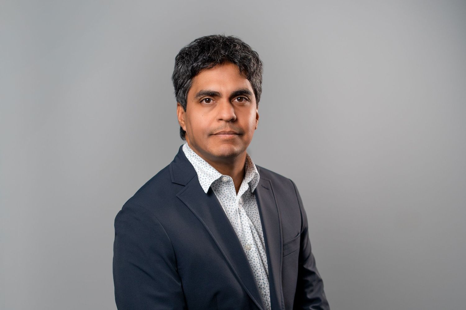 Man in navy blazer posing for professional studio headshot on gray background