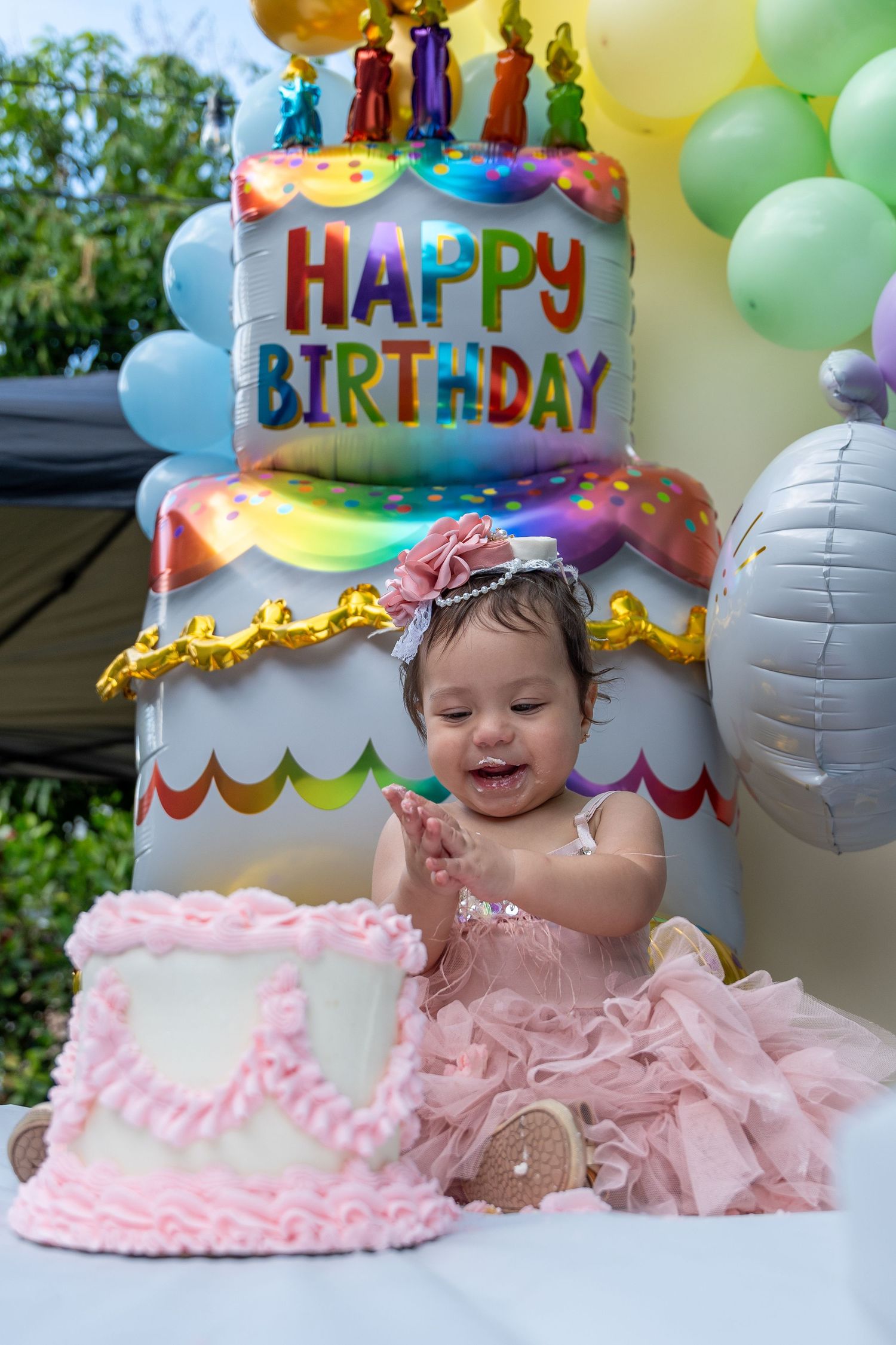 First birthday portrait of a baby girl sitting in front of a colorful birthday cake display during an outdoor celebration.