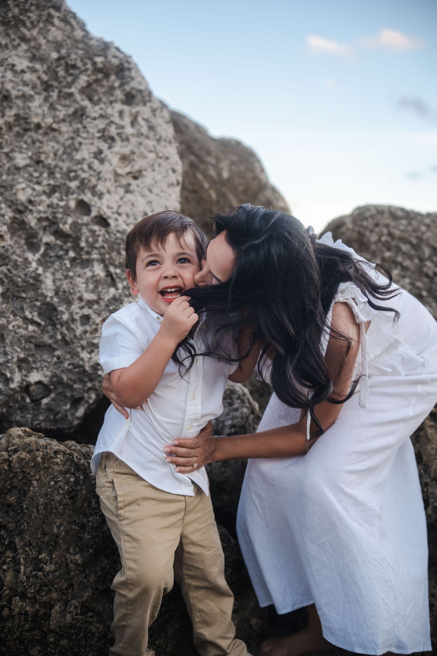 Mother embracing her child near coastal rocks during a pregnancy announcement session.