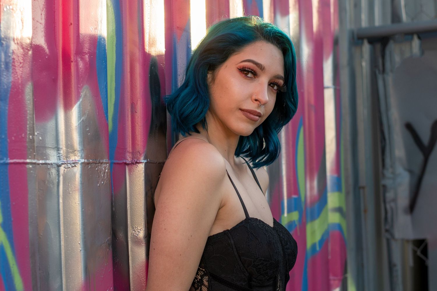 Edgy street-style portrait of a woman with teal hair, leaning against a vibrant graffiti wall in an urban setting. Fort Lauderdale FL