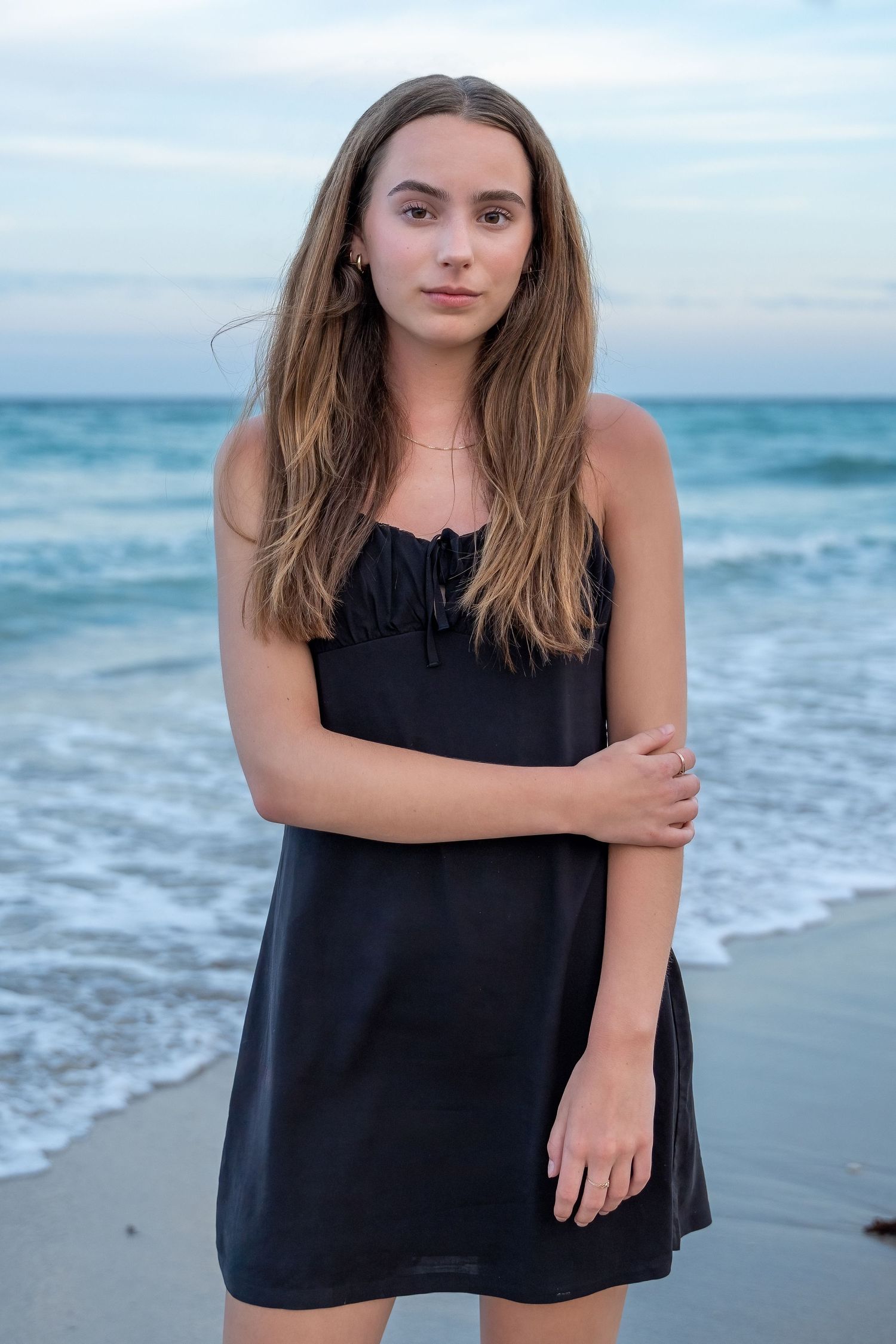 Senior girl in a black dress poses on a Florida beach at sunset, celebrating her graduation with an elegant and natural portrait.