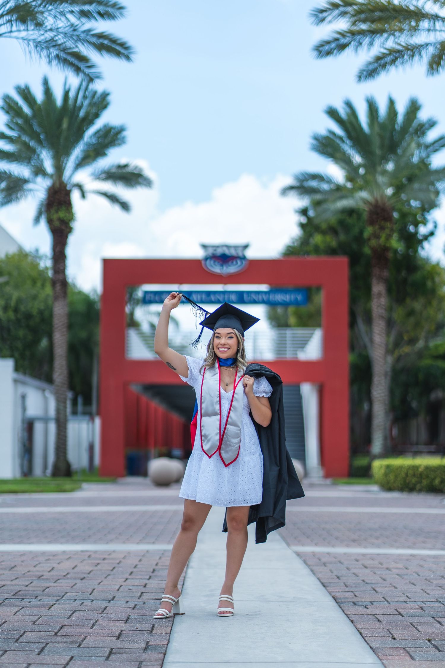 Smiling FAU graduate poses with her cap and stole in front of the red arch at Florida Atlantic University in Boca Raton, FL.