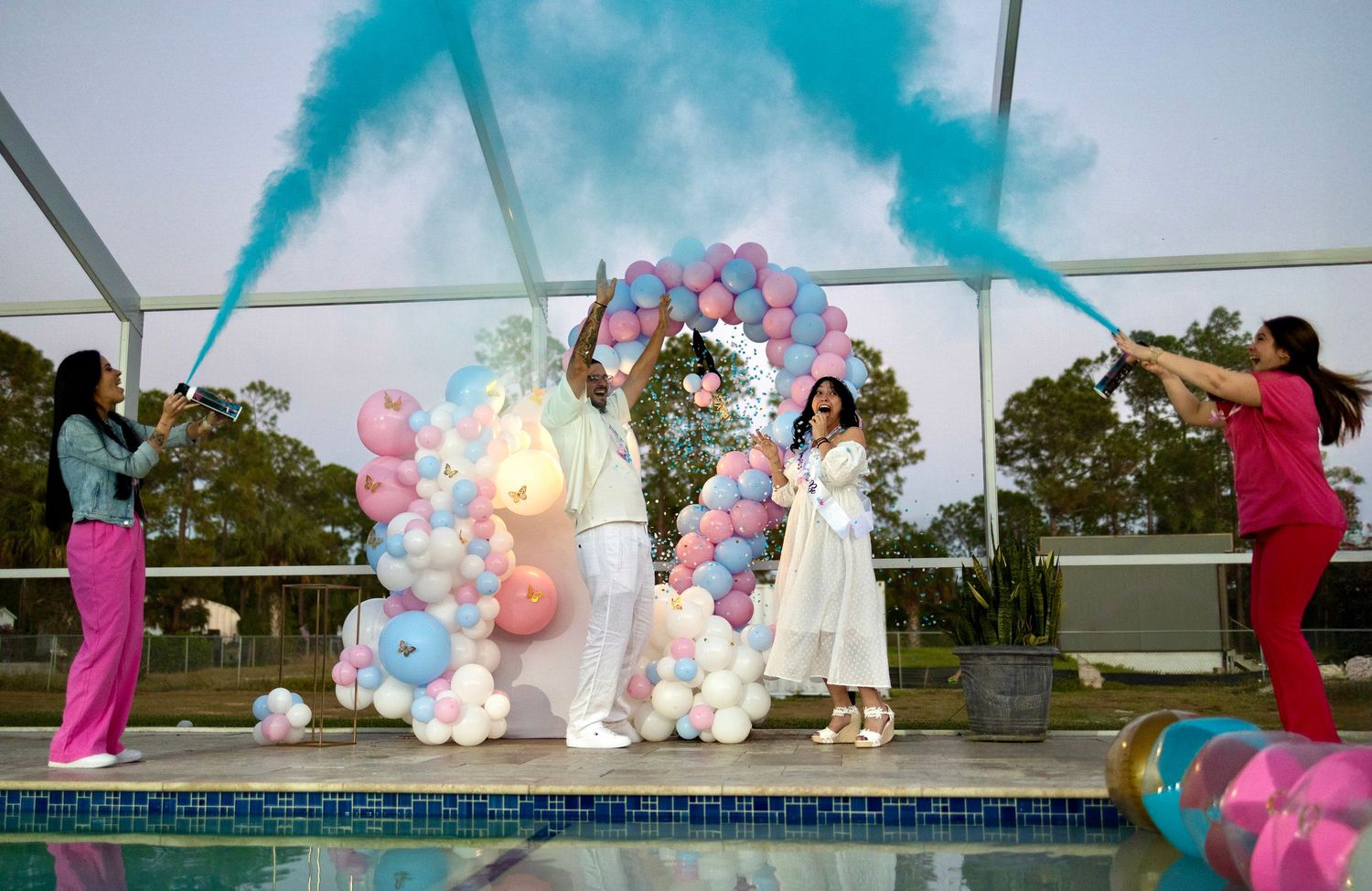 Gender reveal celebration with colored smoke, balloons, and guests reacting during an outdoor event in South Florida.