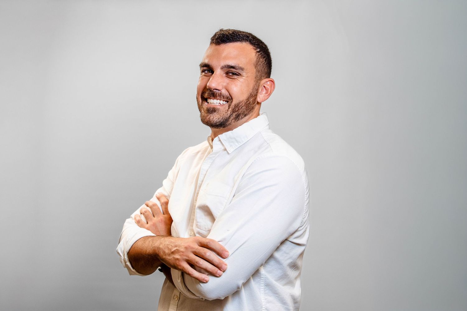 Professional wearing a crisp white button-down shirt stands with arms crossed and a warm smile against gray background.