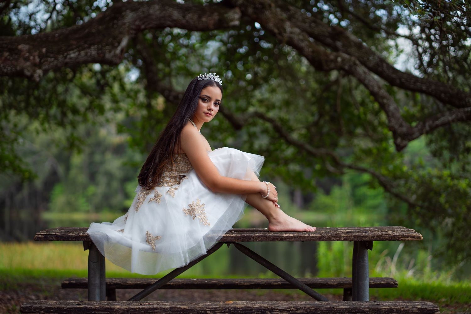 Teen girl in a white tulle dress sitting on a bench under a tree canopy during her birthday shoot.