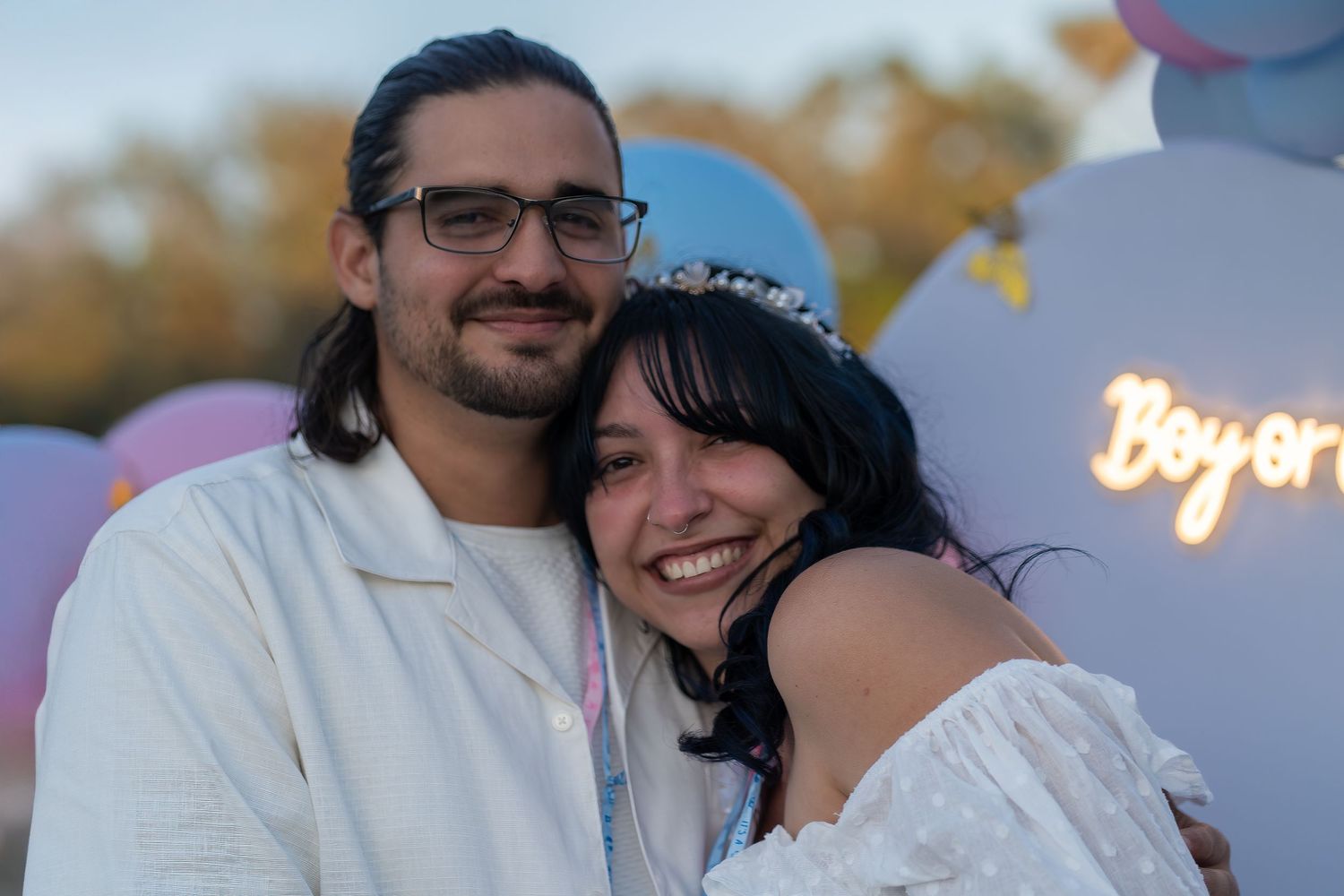 Expecting couple smiling and embracing beneath a pink and blue balloon backdrop at a South Florida gender reveal celebration.