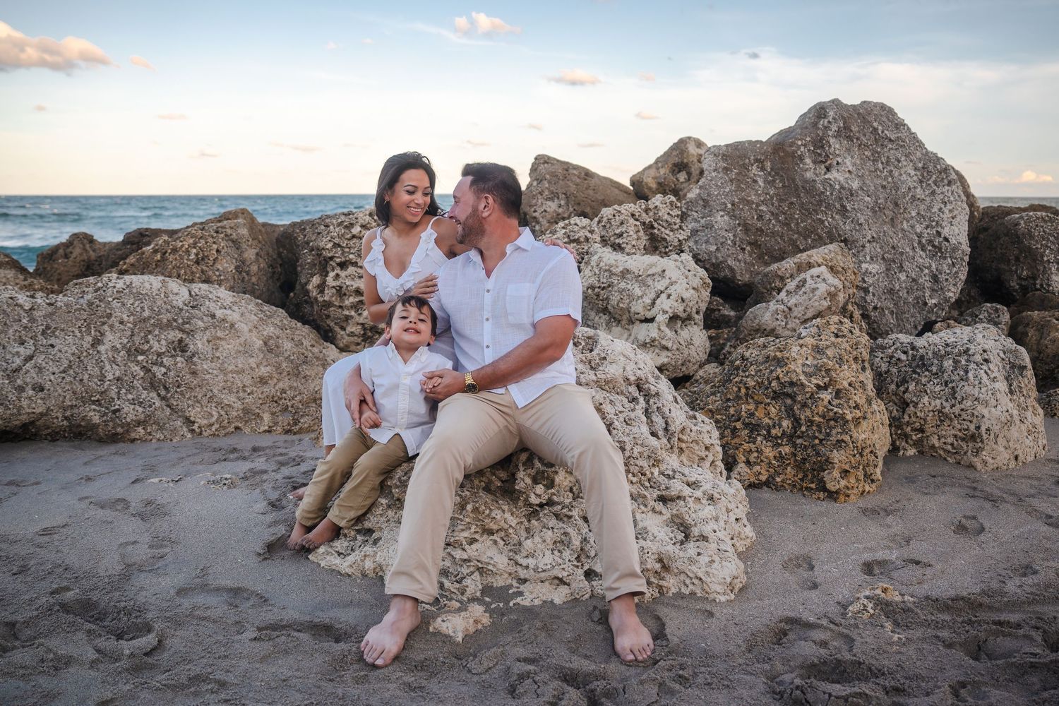 Beach pregnancy announcement photo of an expecting family seated on coastal rocks at sunset in Deerfield Beach, Florida.