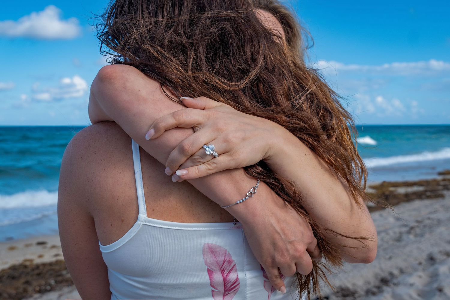 Beach proposal photography capturing a newly engaged couple, with a focus on the engagement ring against the ocean backdrop.