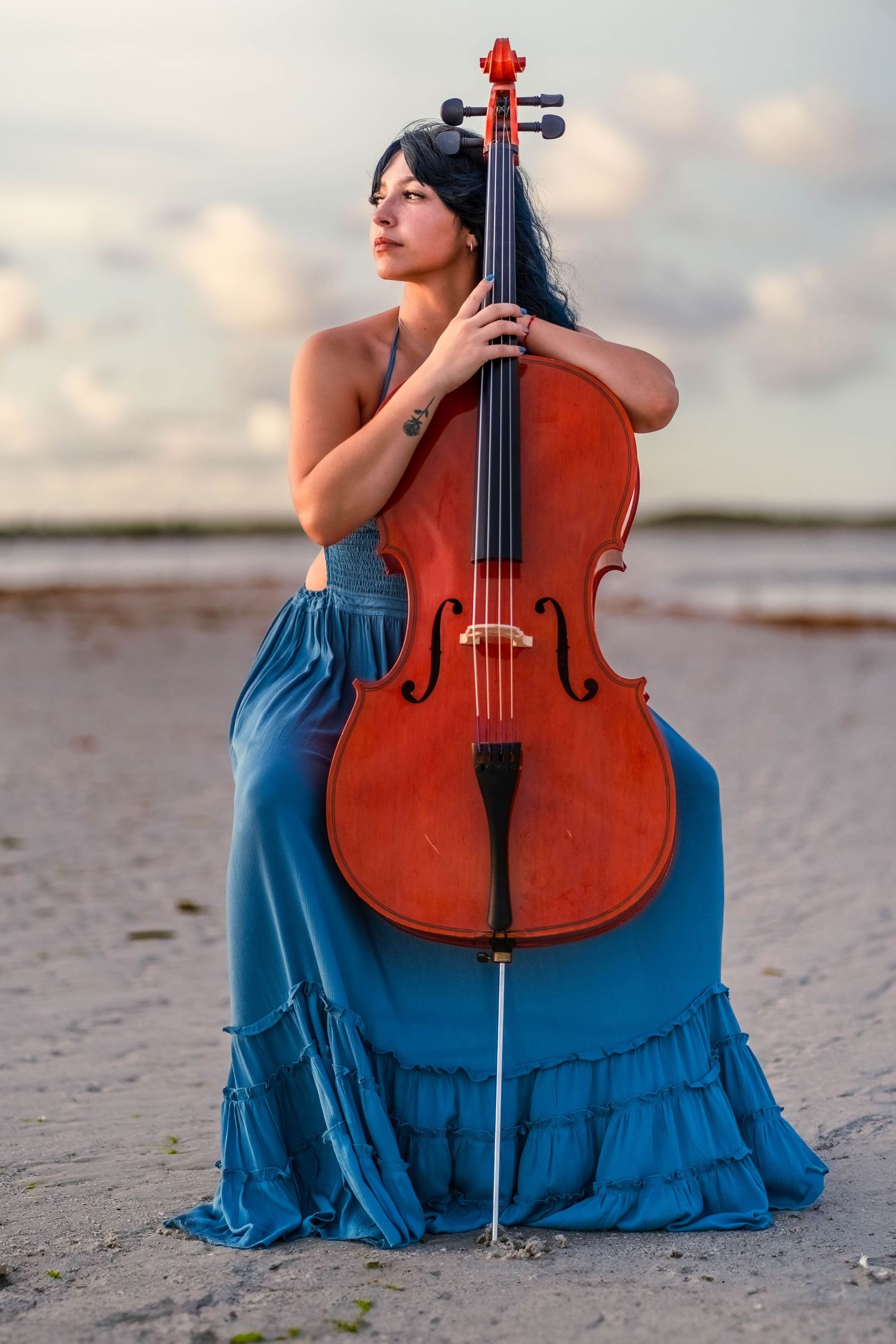 Artistic music-inspired portrait on a South Florida beach – cellist in a blue dress holds her instrument against a dreamy sunset backdrop.