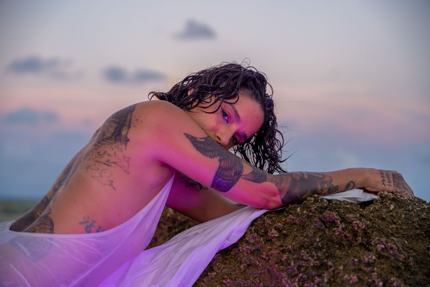 Moody beach portrait of a tattooed man draped in a wet white shirt, lying on the rocks as the golden sunset casts a soft glow.