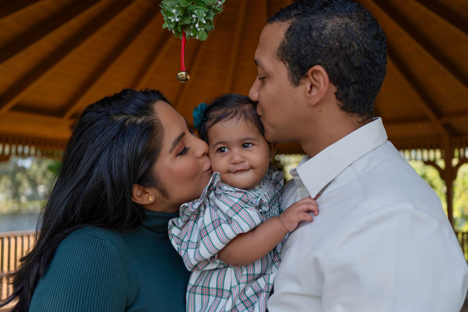 Festive holiday mini session in Miami, FL – parents kissing their baby under mistletoe in a cozy Christmas gazebo setting.