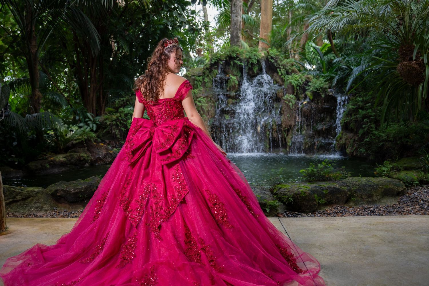 Cinematic quinceañera portrait highlighting a bold burgundy gown set within a tropical orchid garden.