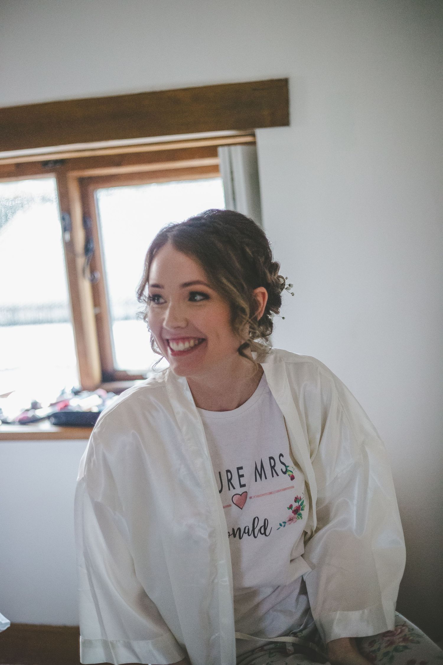 Bride in white robe smiling while getting ready in a room with natural window light.
