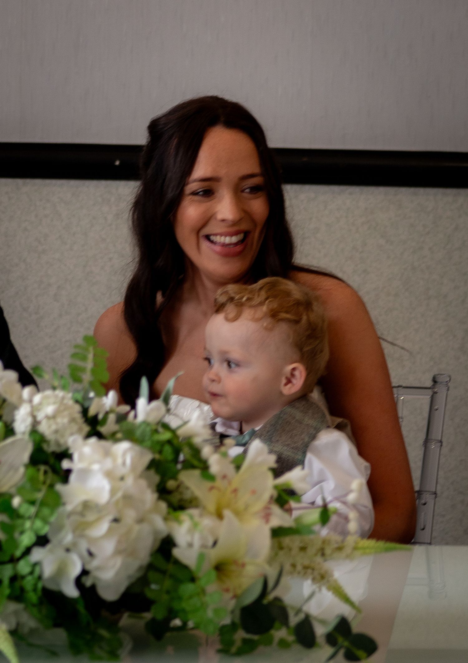 A woman and toddler sit at a table with elegant white floral arrangements at what appears to be a formal event.