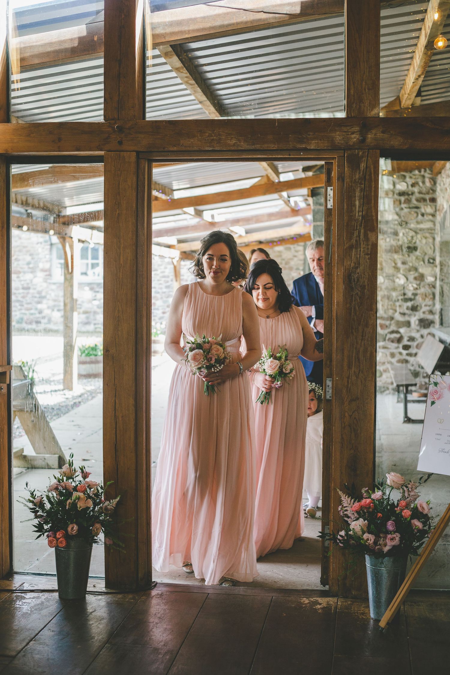 Bridesmaids in flowing pink dresses stand in a rustic venue with stone walls and wooden beams.