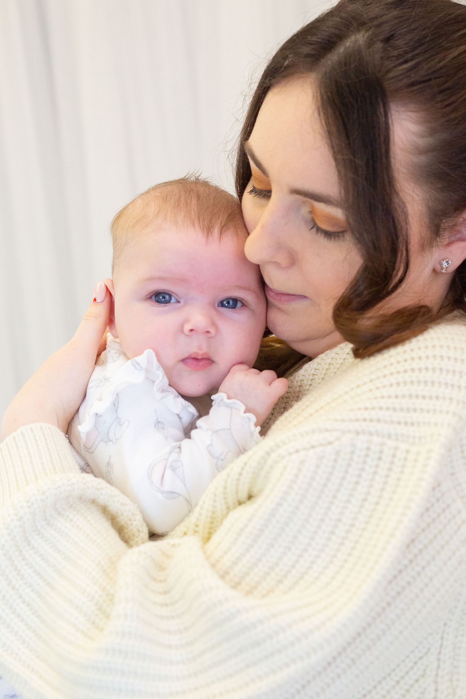 Tender moment between mother and baby in white outfit against light background.