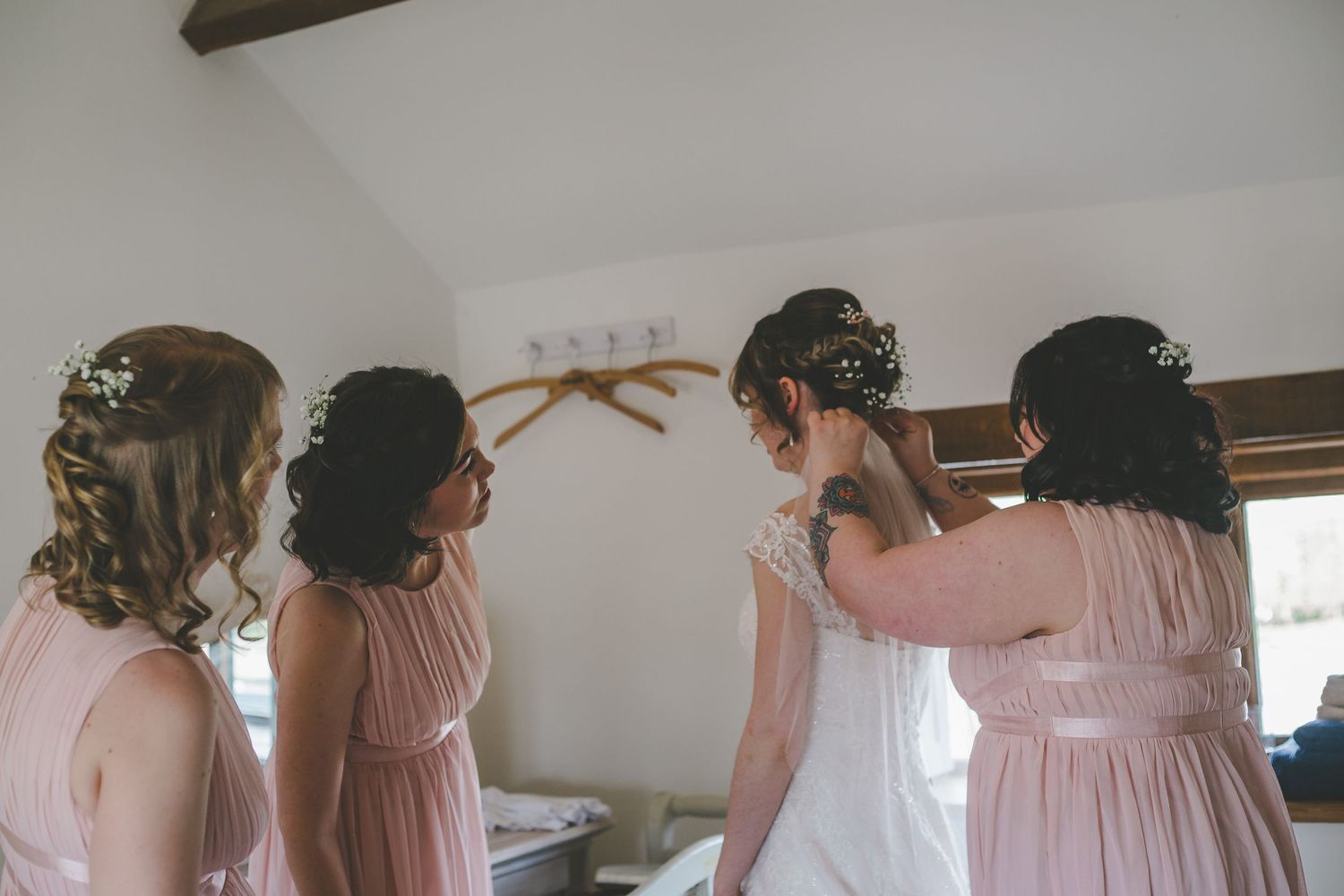 A group of bridesmaids in blush pink dresses help each other get ready for a wedding ceremony.