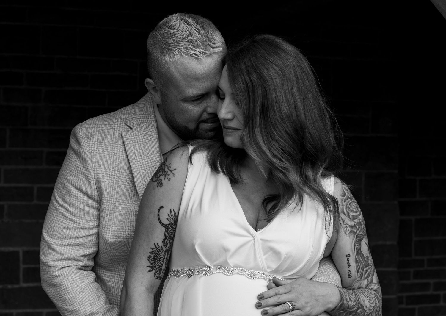 A romantic black and white photo series shows a couple embracing in formal attire against a brick wall.