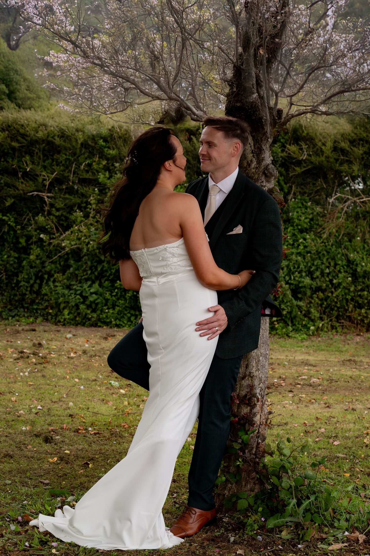 A couple shares a romantic moment outdoors against a tree in formal wedding attire.