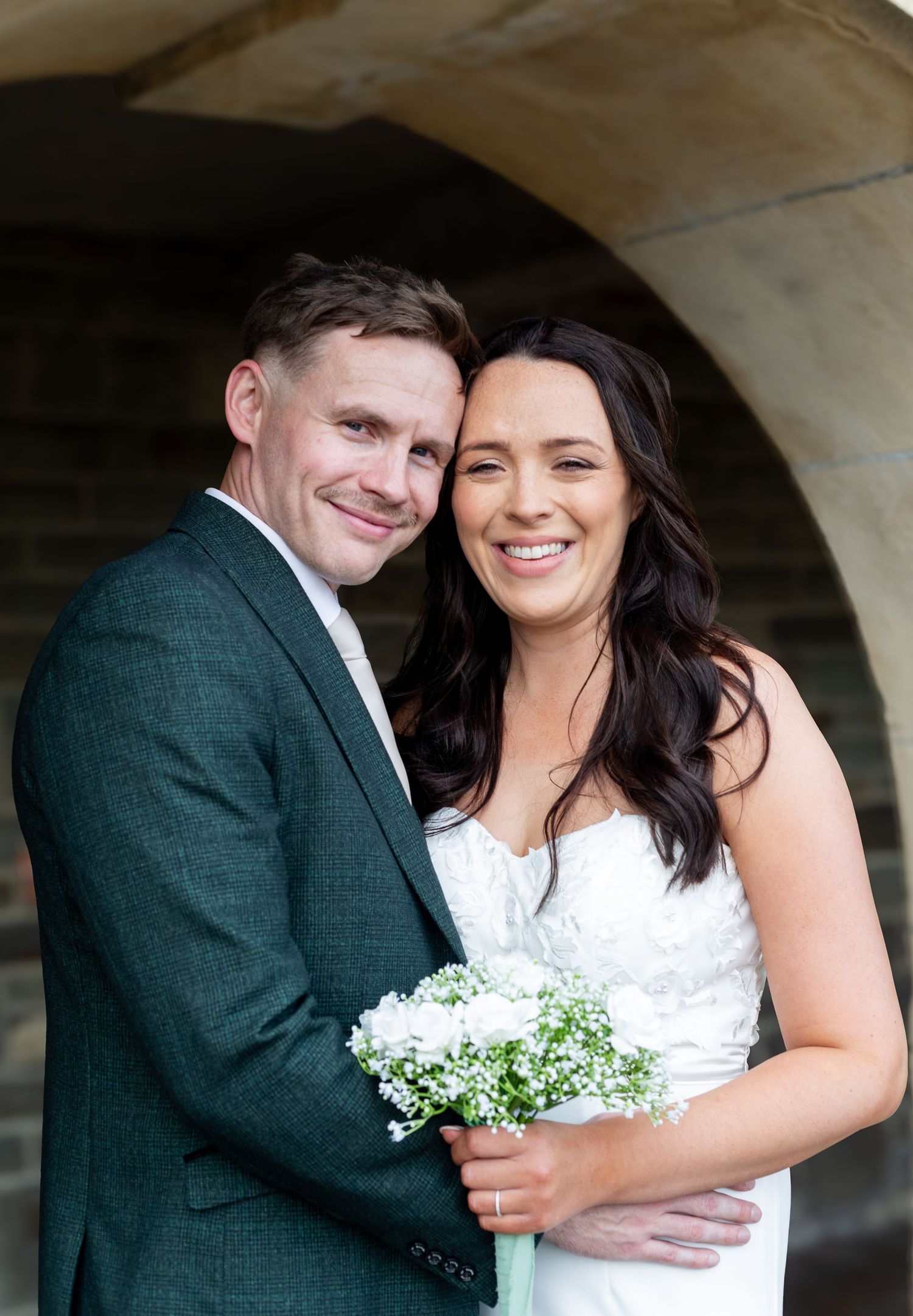 A romantic wedding couple shares tender moments under stone archways with white baby's breath bouquet details.