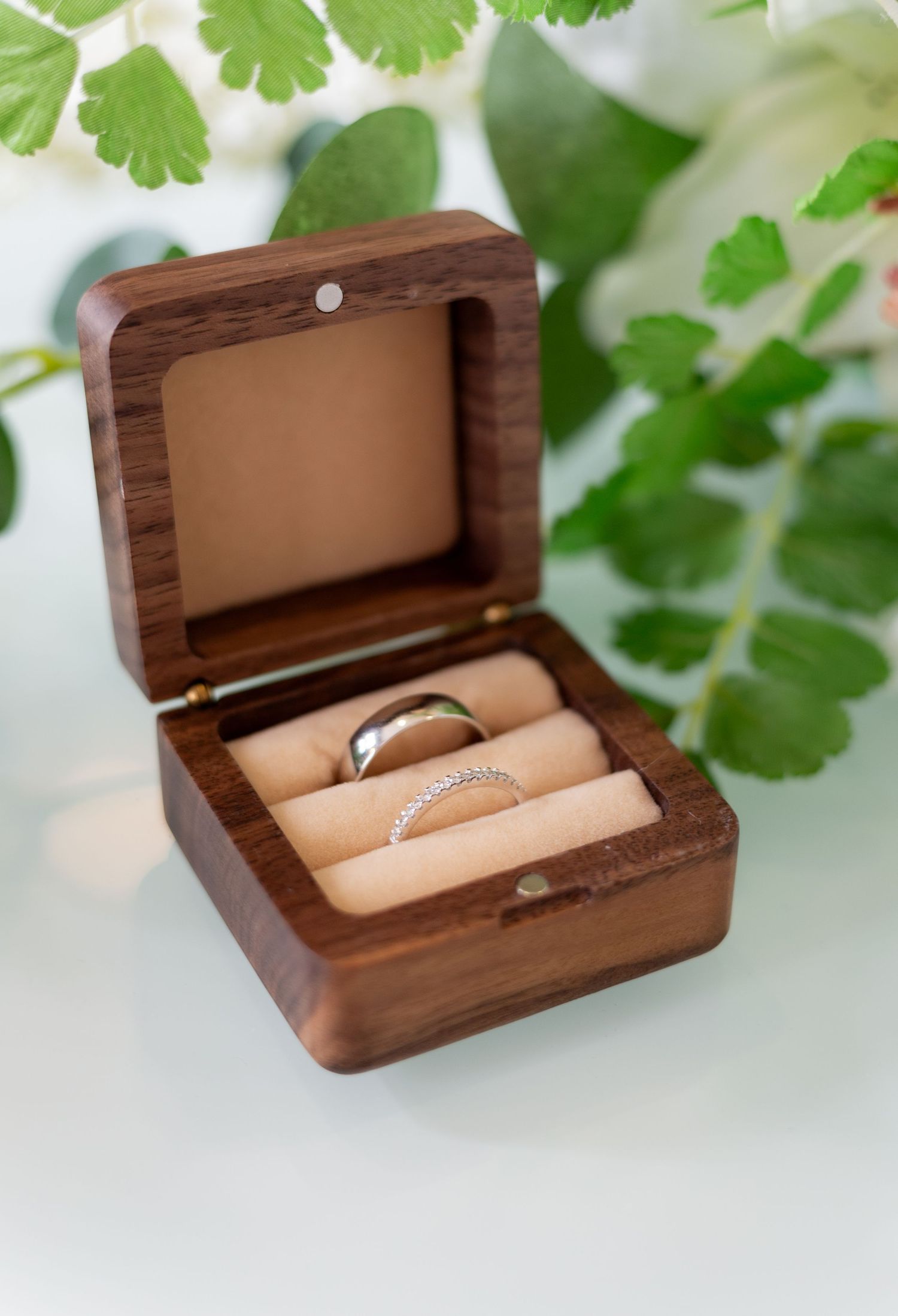 Wooden ring box with hinged lid displaying delicate jewelry against green foliage background.