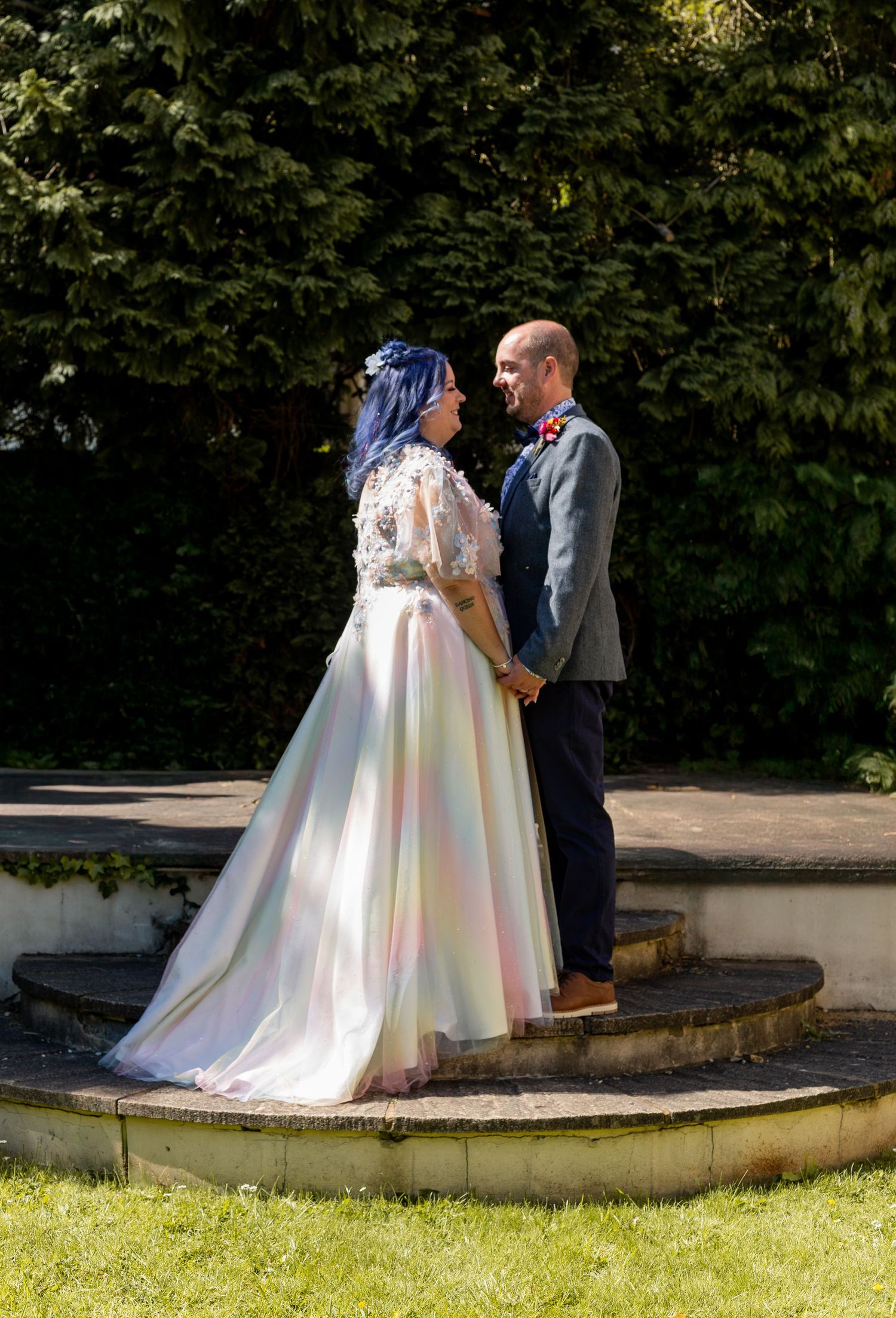 A wedding couple stands on a curved stone path surrounded by lush greenery on a sunny day.