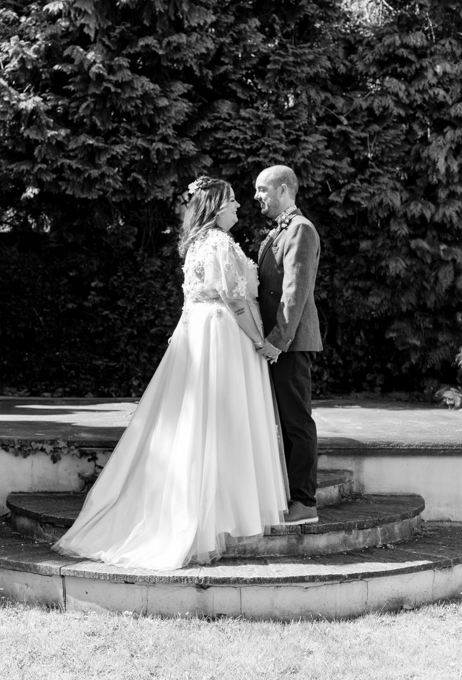 A couple in wedding attire stands together on curved stone steps in a garden setting, captured in black and white.