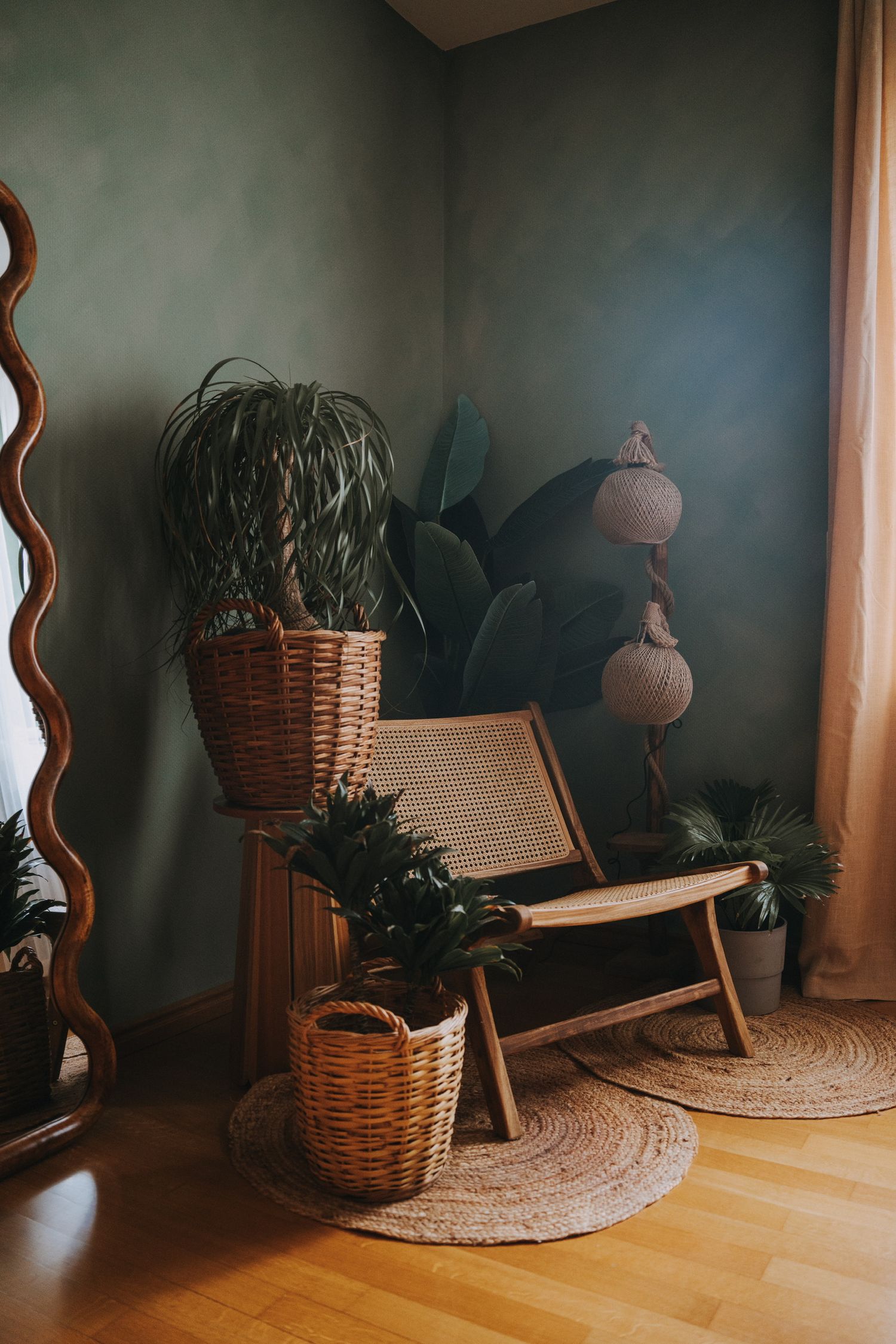 Bohemian corner with rattan chair, wicker baskets, tropical plants, jute rug, and decorative lamp against dark green wall.