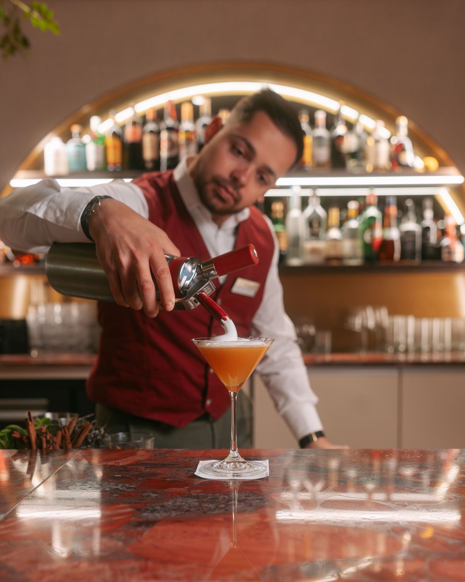 Bartender pouring a frothy orange cocktail into a martini glass at a well-stocked bar.