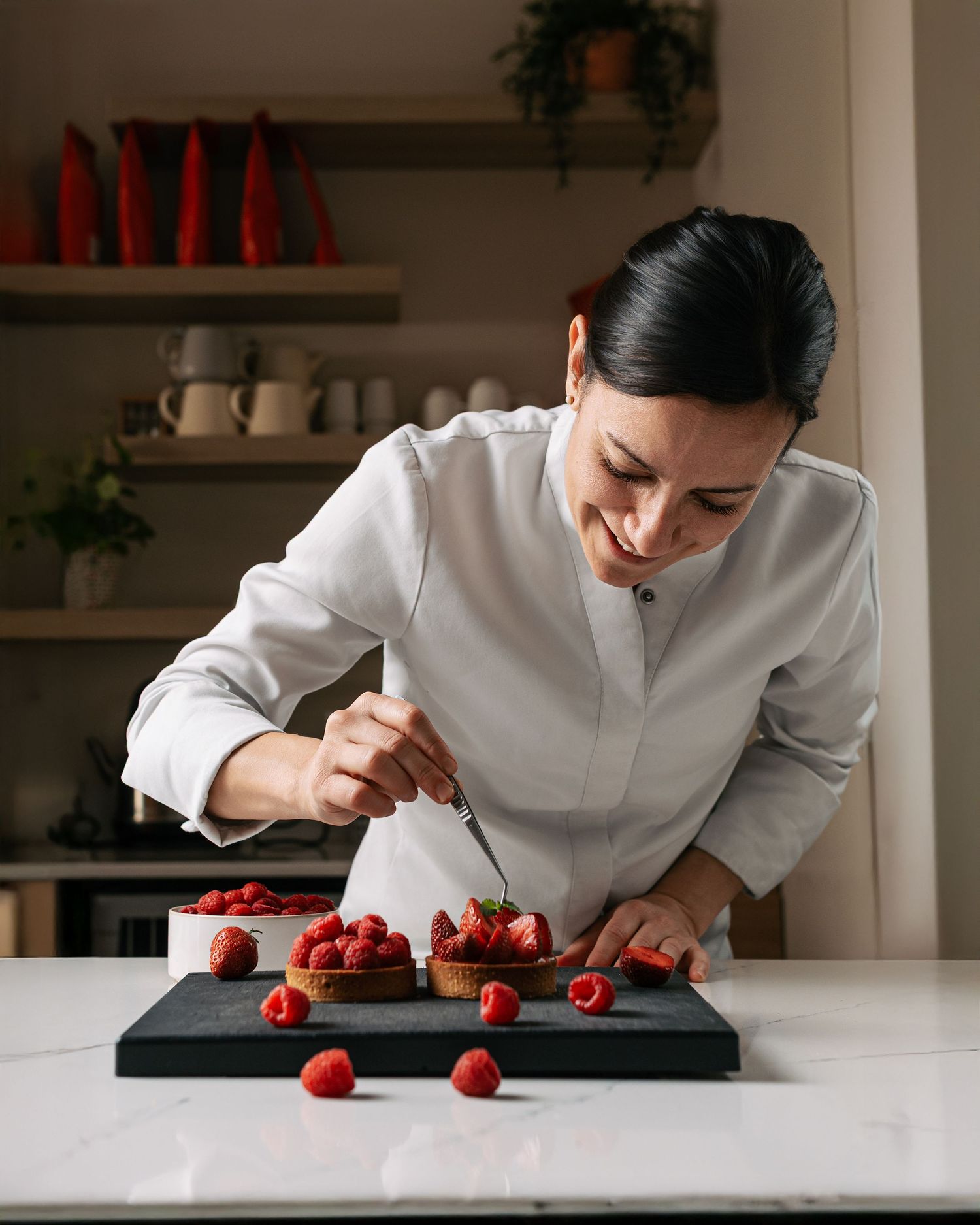Smiling female pastry chef carefully decorating berry tarts on a marble countertop.