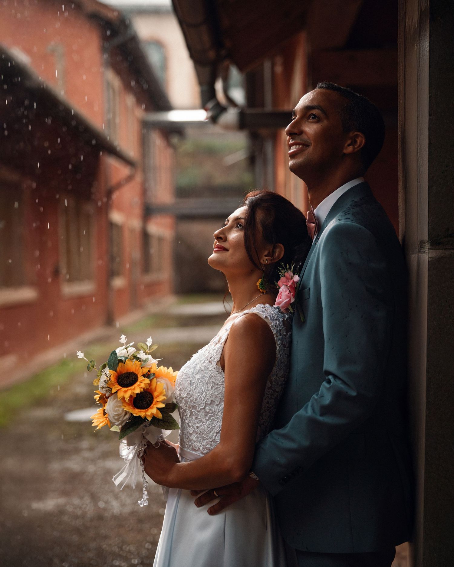 Bride holding sunflower bouquet and groom in teal suit gaze upward together in the rain.