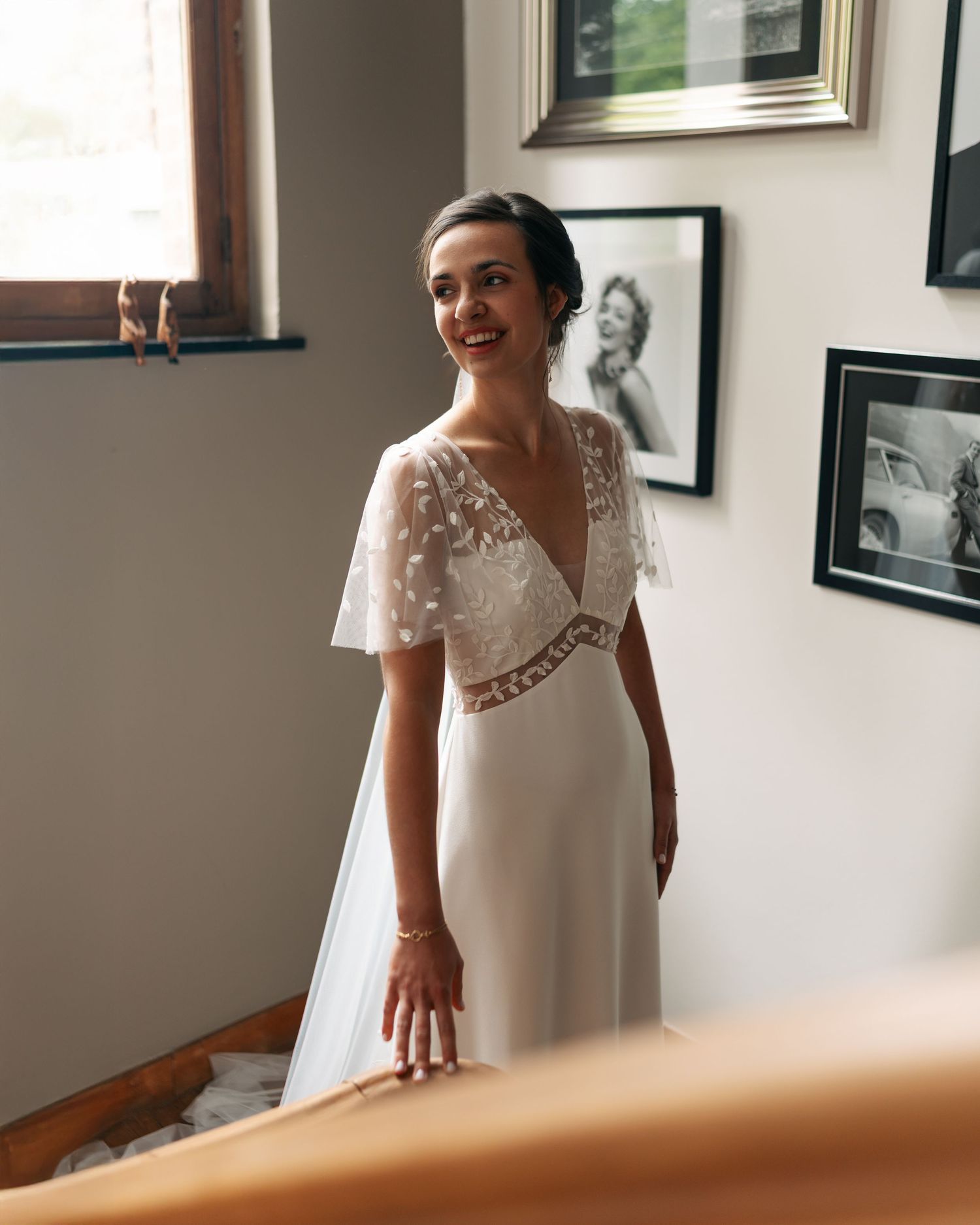 Bride smiles in elegant white V-neck lace wedding dress with flutter sleeves in a sunlit room.