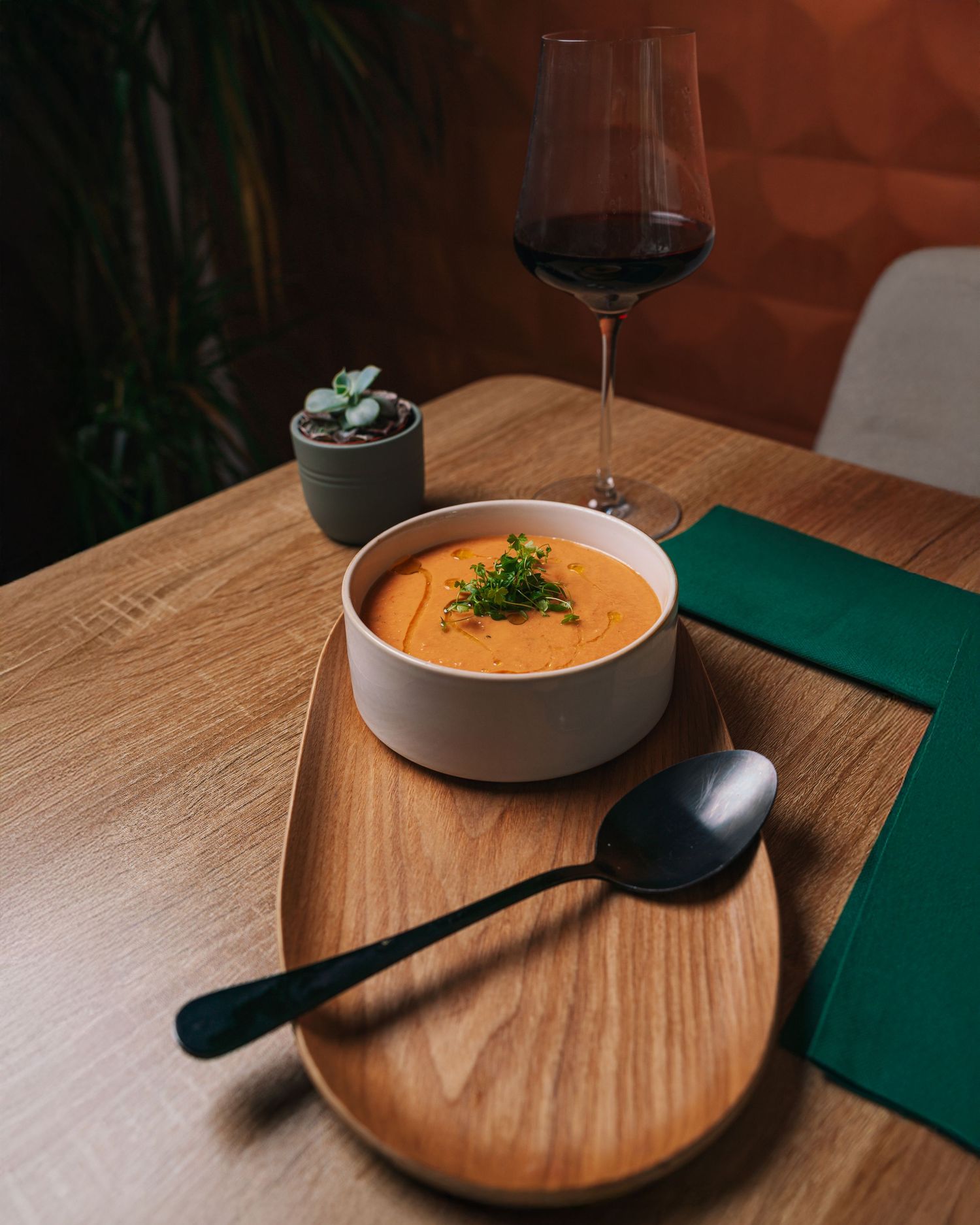 Bowl of vibrant orange cream soup with microgreens on wooden tray next to a red wine glass.