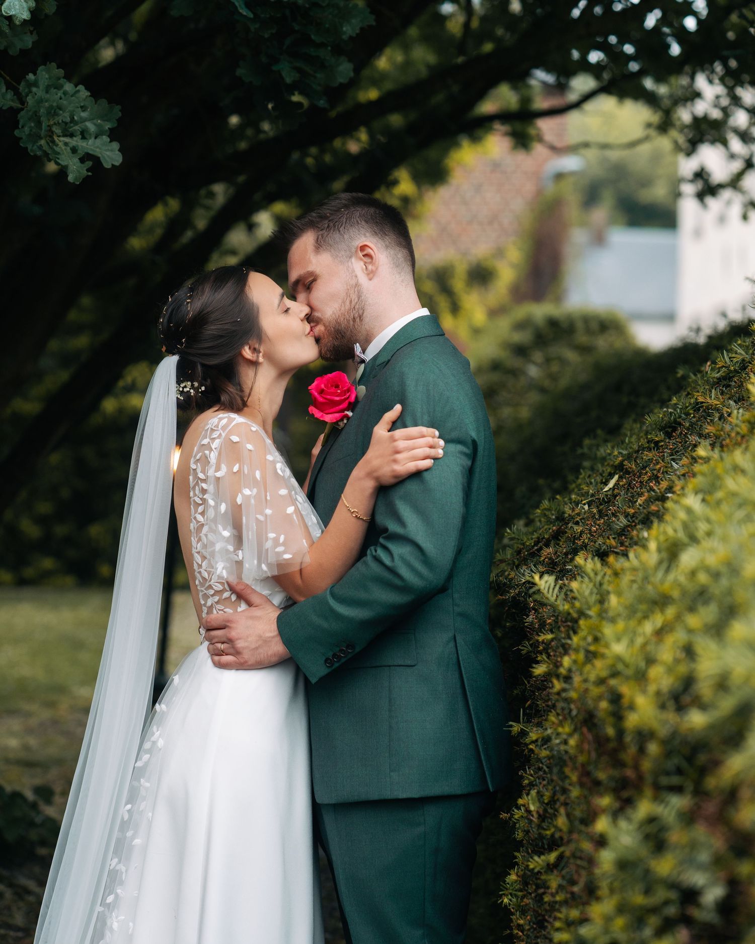 Bride and groom sharing a romantic kiss outdoors, bride in lace veil and groom in green suit with red rose boutonniere.