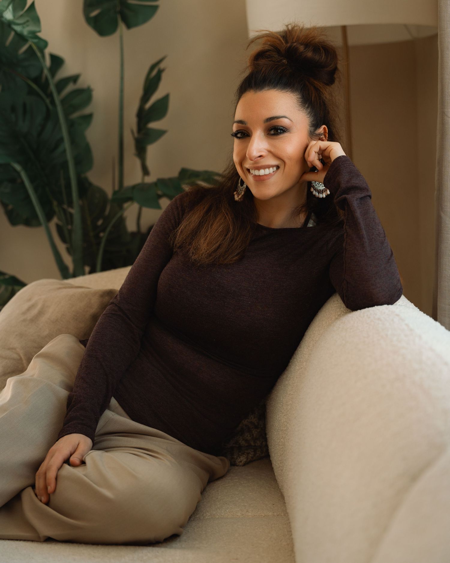 Smiling woman in brown sweater sitting on cream sofa with tropical plant background.