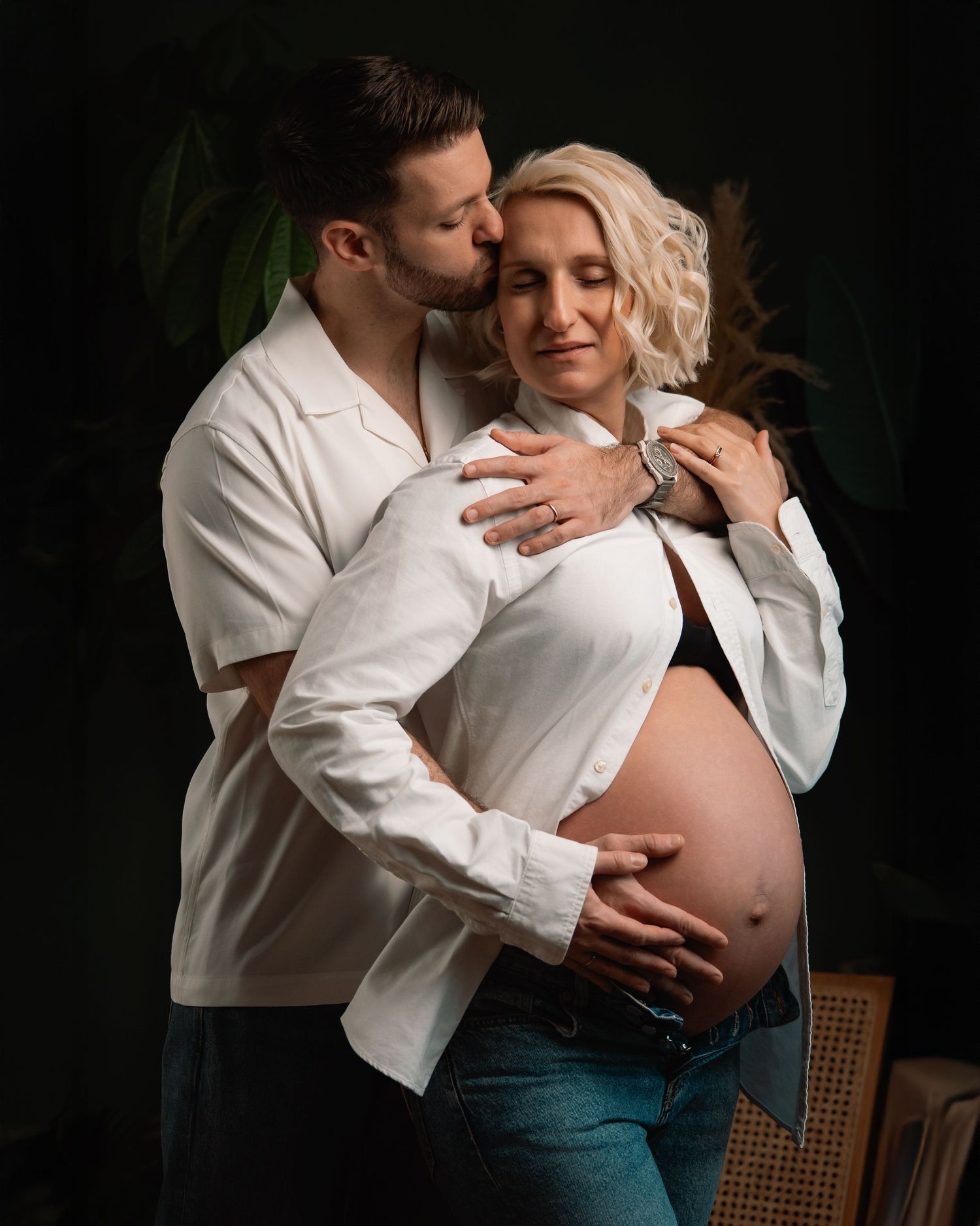 Couple embracing during maternity photoshoot, man kissing pregnant woman's head in studio.