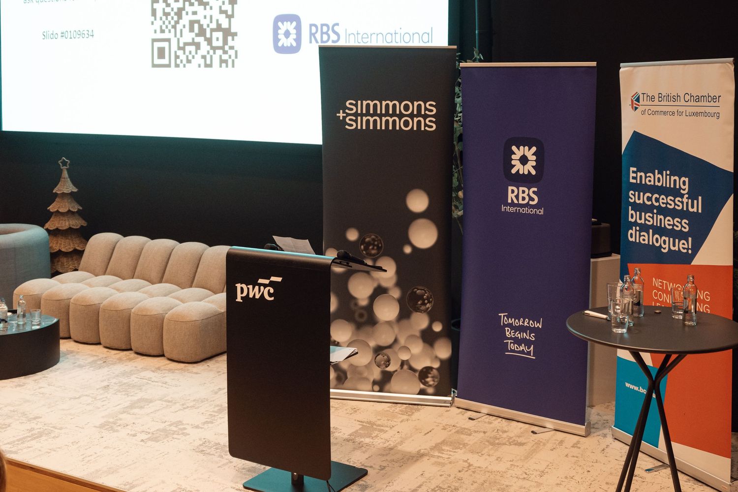 Conference room setup featuring PwC, Simmons and Simmons, RBS International, and British Chamber of Commerce branded banners.