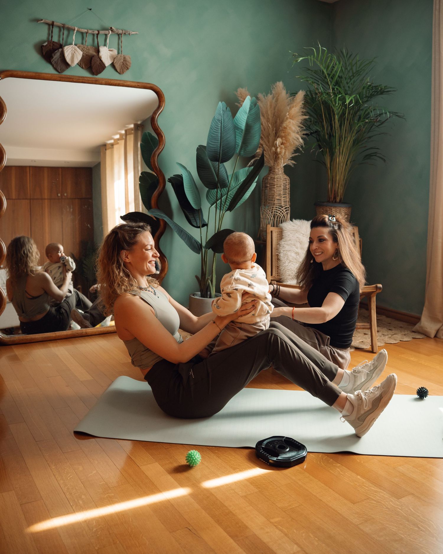 Two women exercise on a yoga mat with a baby in a cozy green-walled home gym.