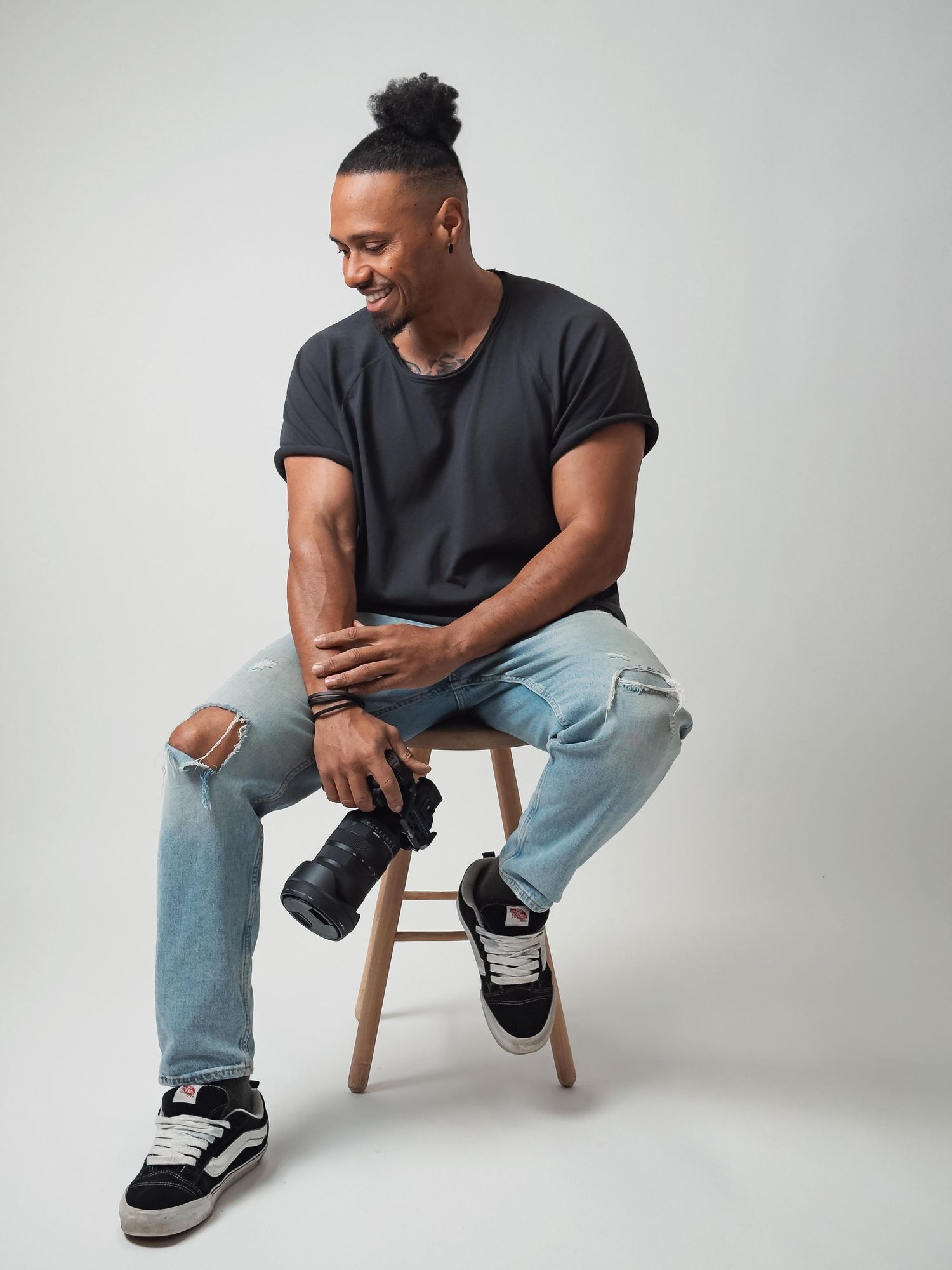Smiling man sitting on stool holding camera wearing black tee and ripped jeans.