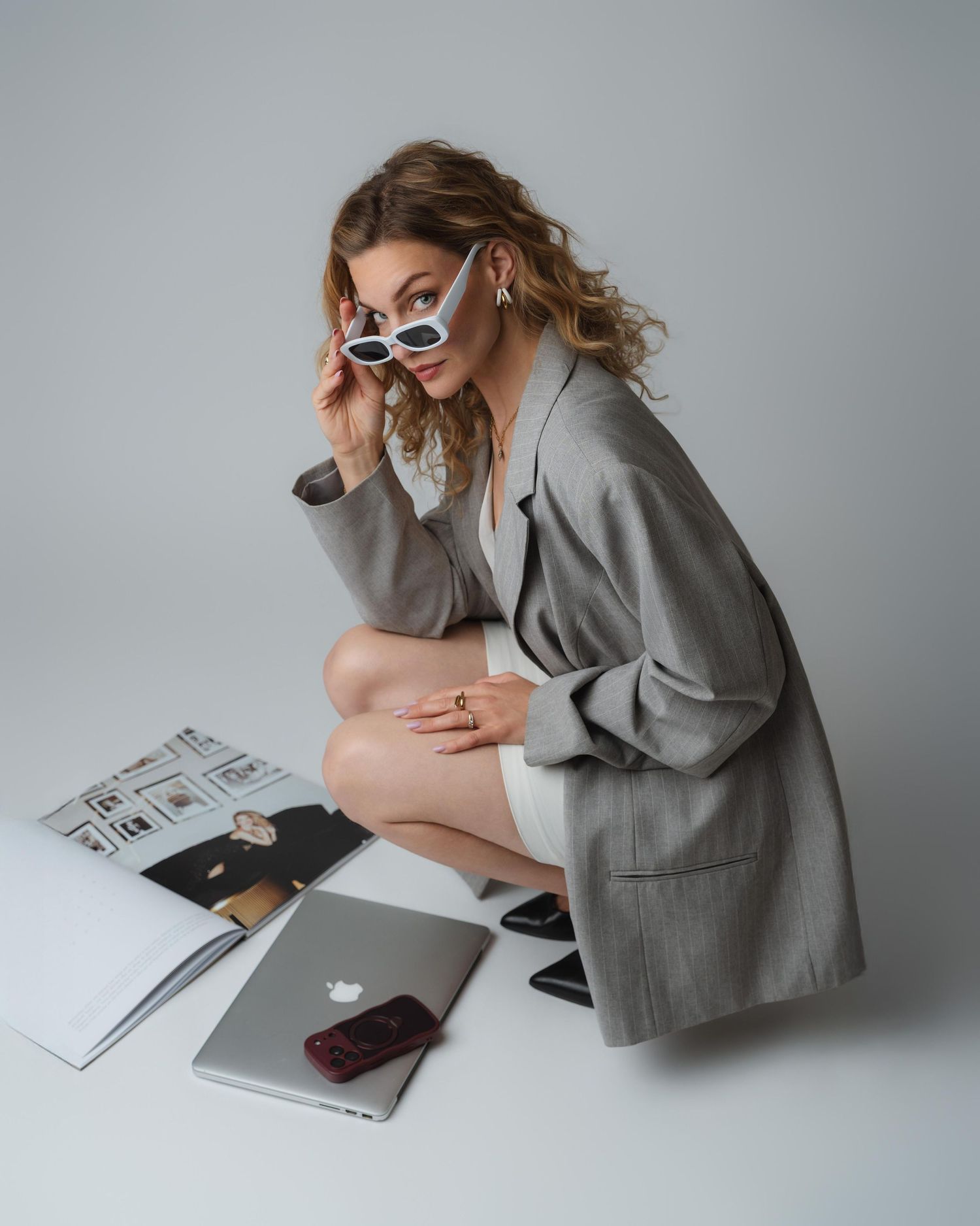 Woman in gray blazer and white sunglasses sits on floor with MacBook, magazine, and phone in a studio setting.