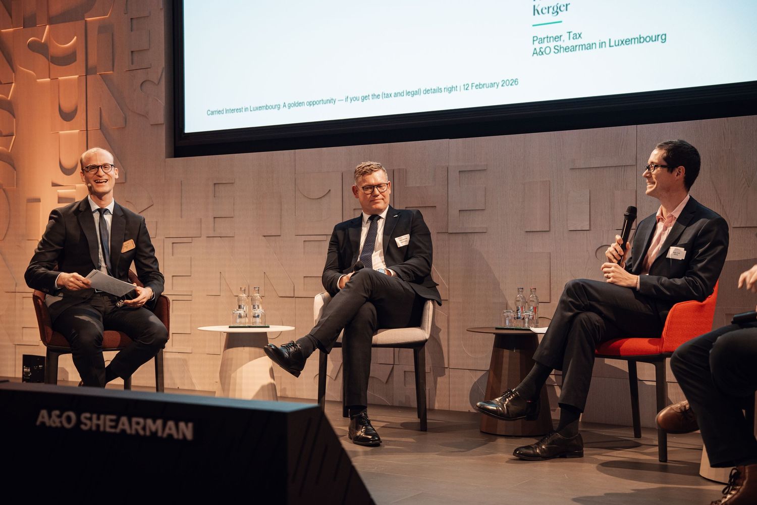 Three business professionals in discussion on stage at A&O Shearman panel event.