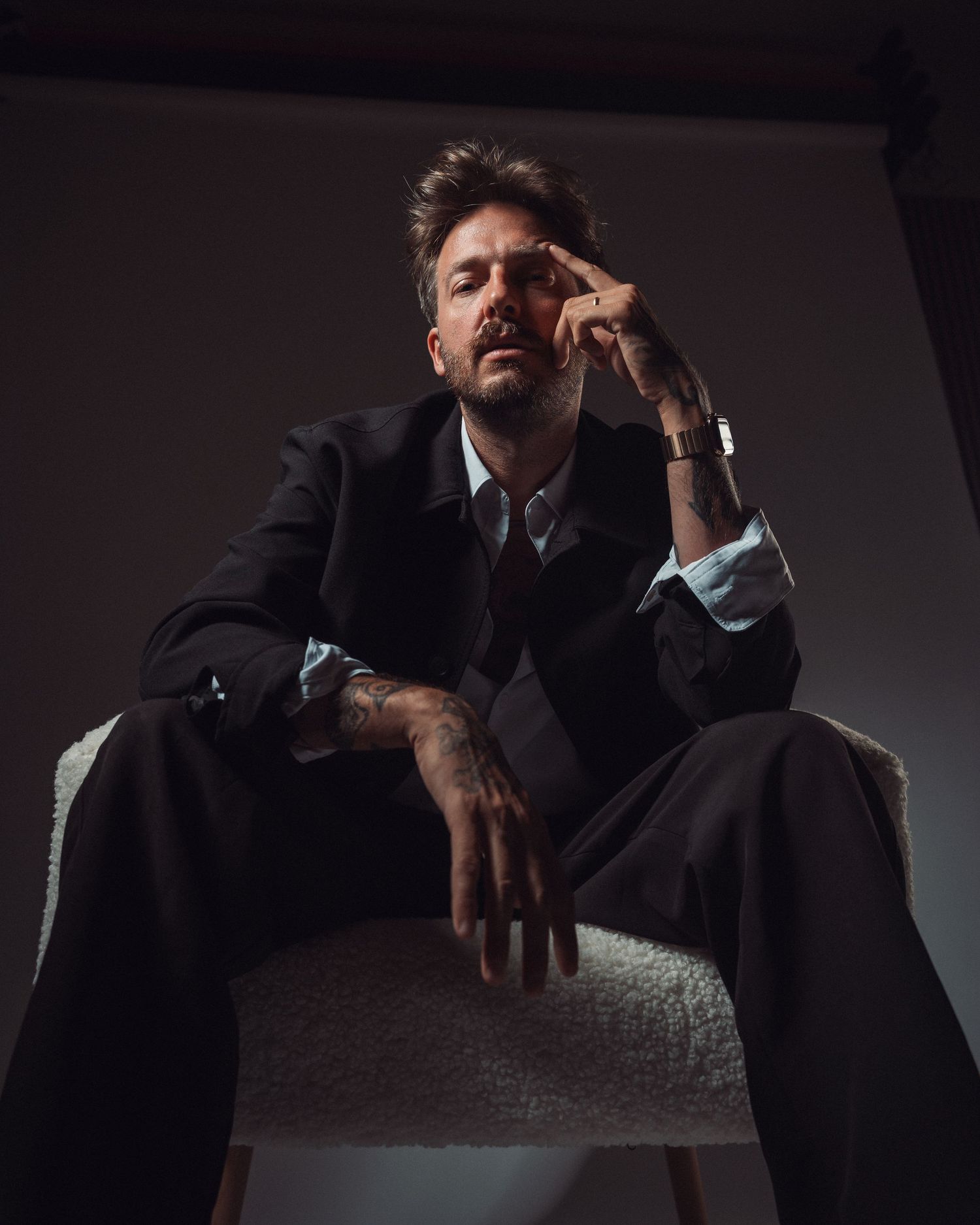Tattooed man in dark suit sitting on white chair, posing with hand near face in moody studio portrait.
