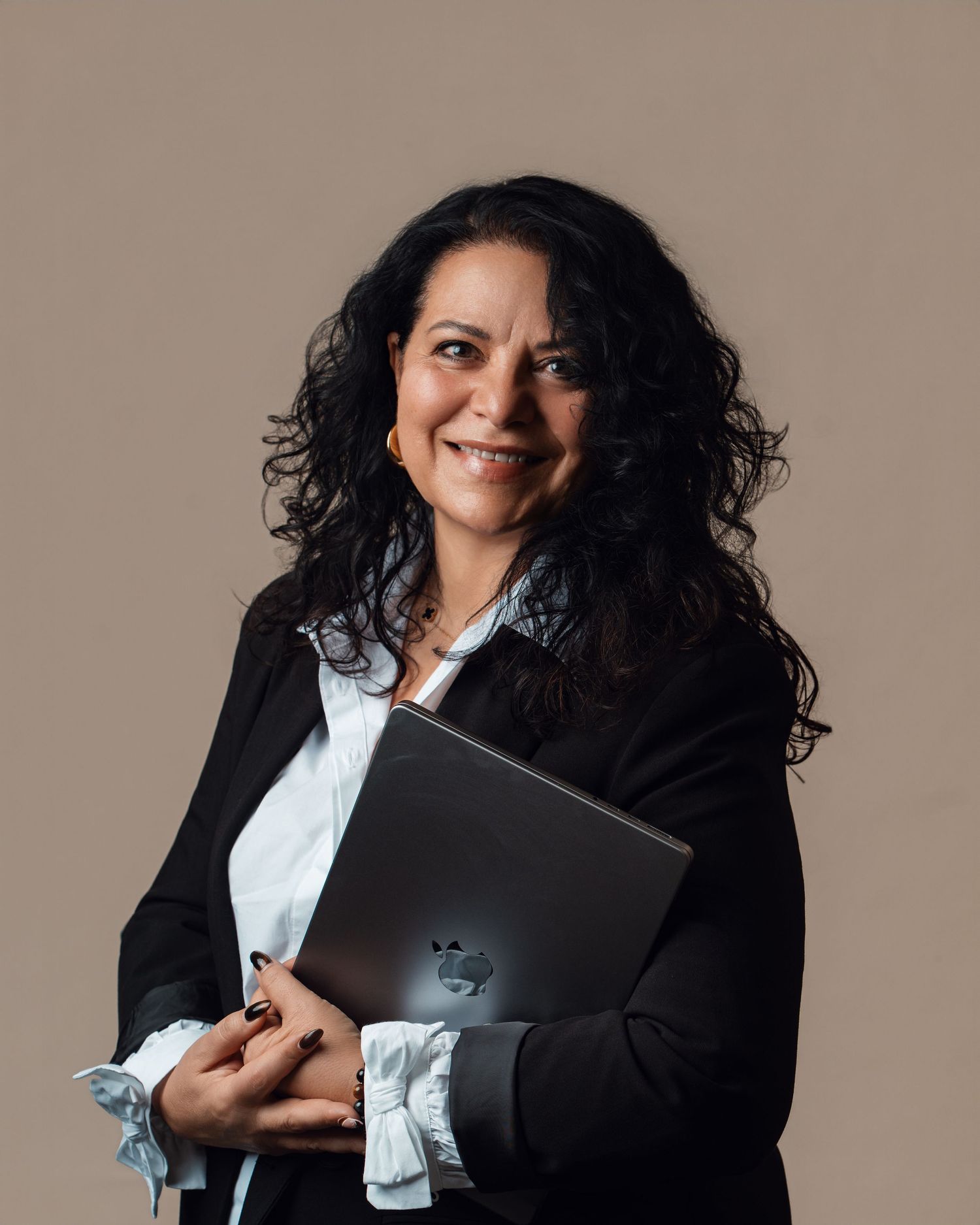 Cheerful curly-haired woman in a black blazer holding a laptop against a warm beige background.