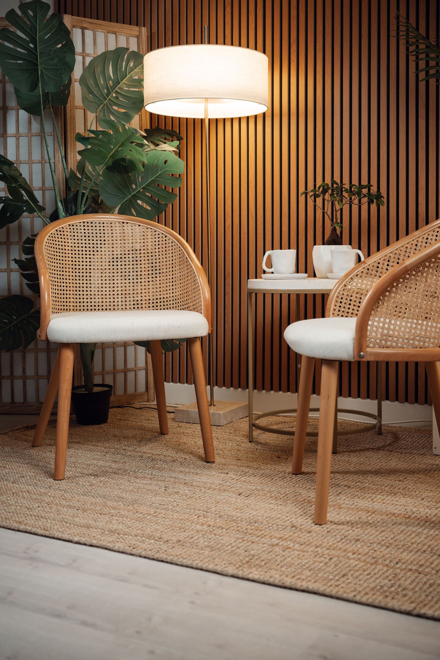 Two rattan chairs with white cushions on a jute rug, beside a floor lamp and small table in a warm modern interior.