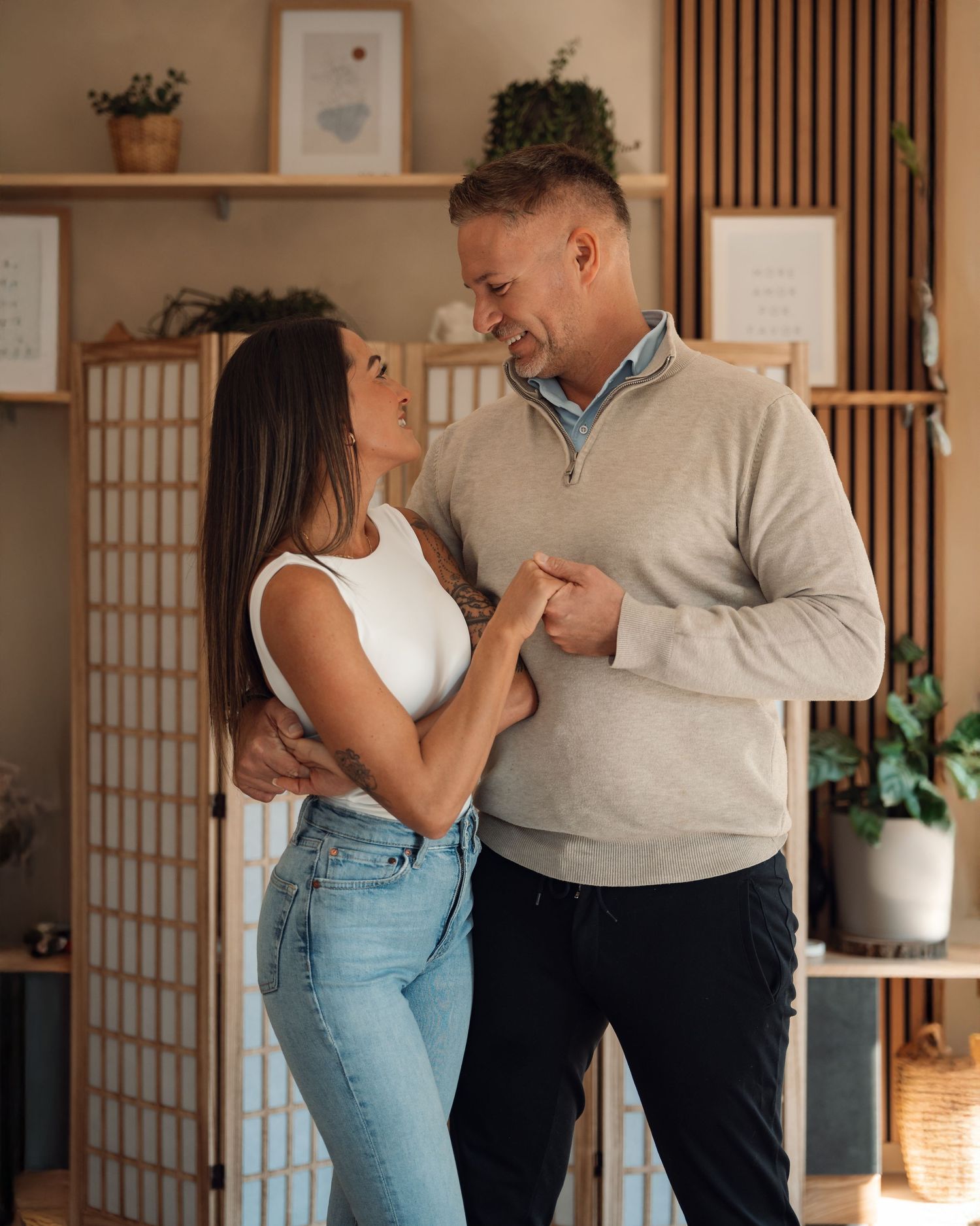 A couple smiles at each other while holding hands in a cozy modern living room with wooden decor.
