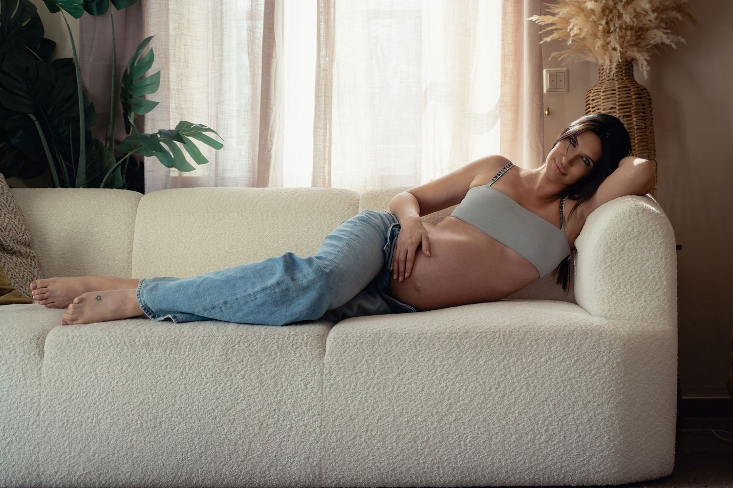 Pregnant woman in jeans relaxes on a white boucle sofa in a bright living room.