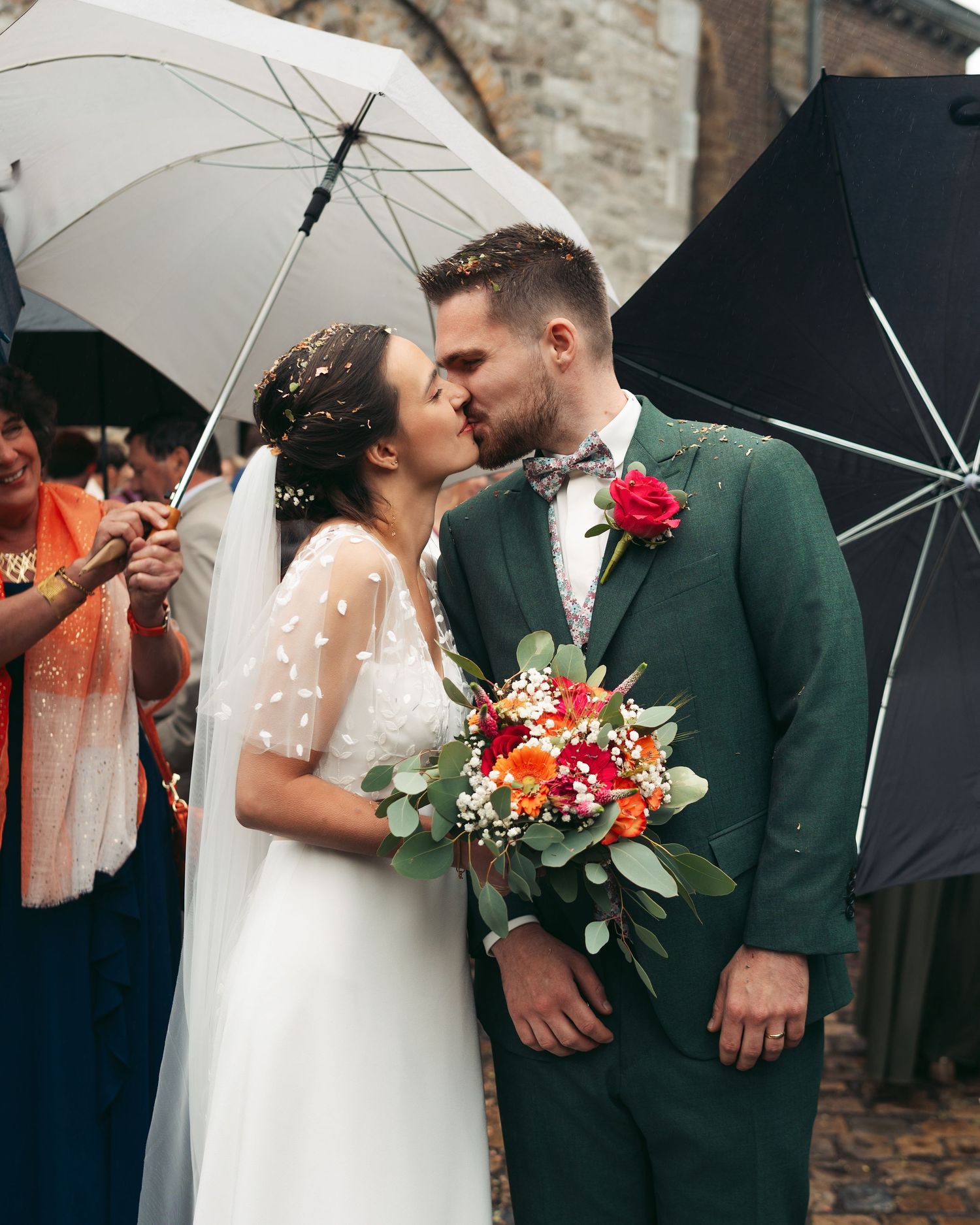 Bride and groom share a first kiss under umbrellas with colorful confetti at wedding ceremony.