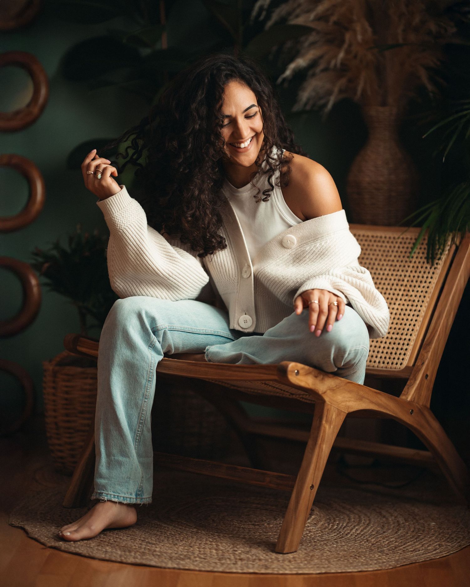 Smiling curly-haired woman in white cardigan and light jeans sitting on wooden rattan chair in boho studio.
