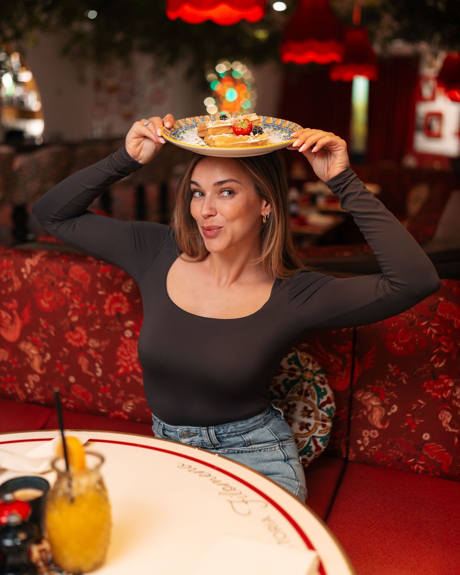 Playful woman balancing a plate of pancakes on her head at a vibrant red restaurant booth.
